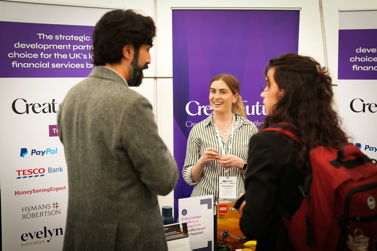 A smiling woman with a lanyard interacting with a man in a tweed jacket and a woman with a red backpack at a "CreateFuture" exhibition stand