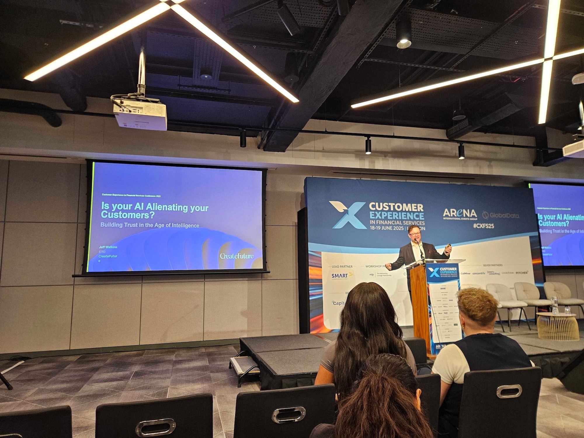  A wide shot captures a conference presentation from the audience. A speaker in a suit stands at a podium on a stage, addressing an audience, with 