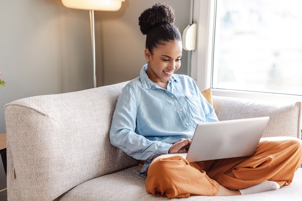 A woman sitting cross-legged on a couch, focused on her laptop.