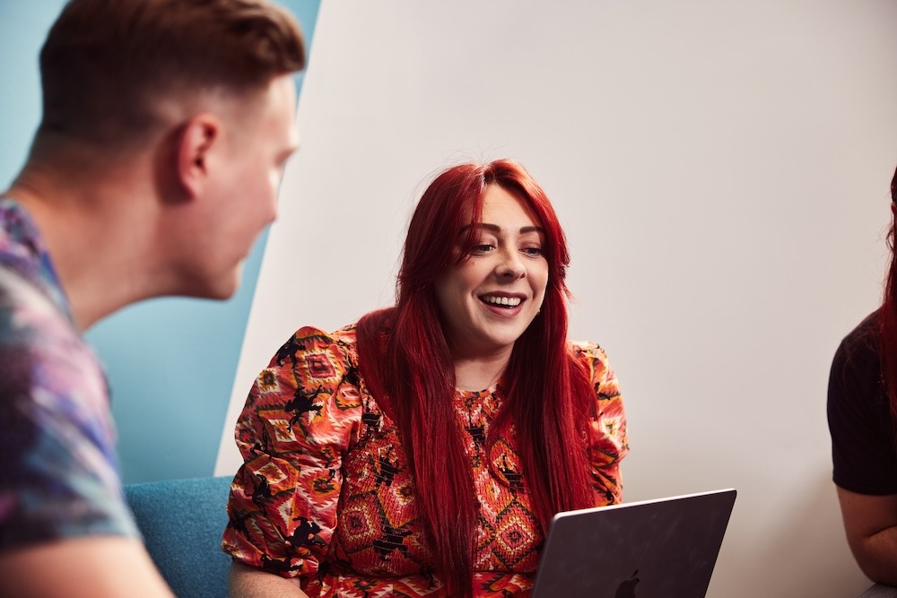 A woman with long red hair, wearing a colourful patterned top, smiles while looking at a laptop during a conversation with two other people.