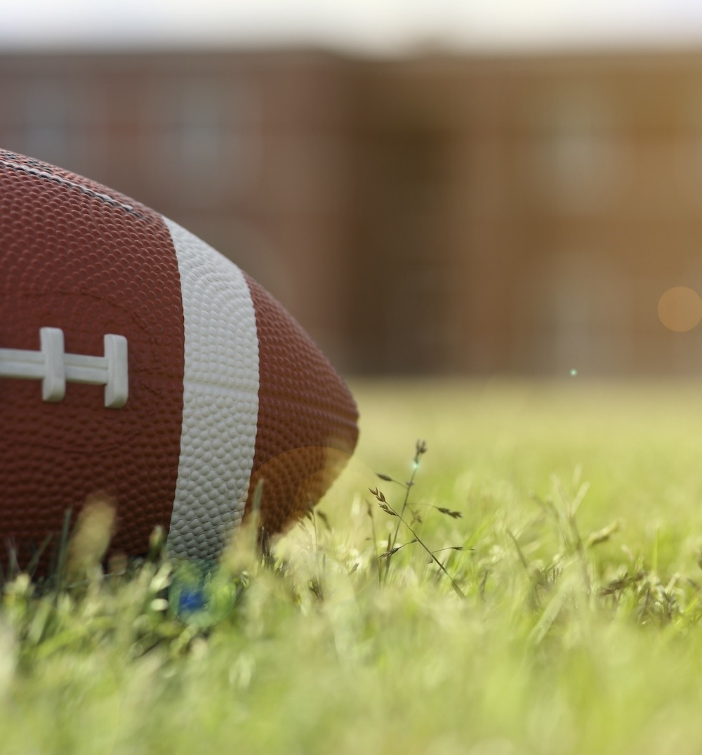 Close-up of an American football resting on a grassy field with a blurred building in the background.