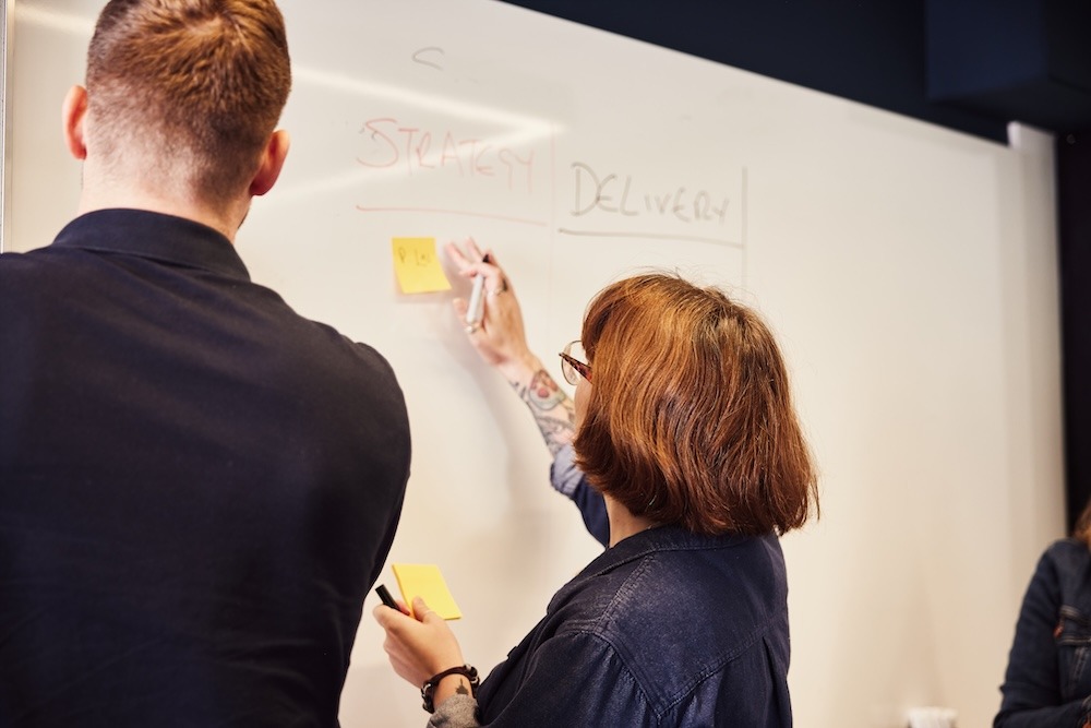 Two people collaborating at a whiteboard during a meeting. One person is putting a yellow sticky note on the board.