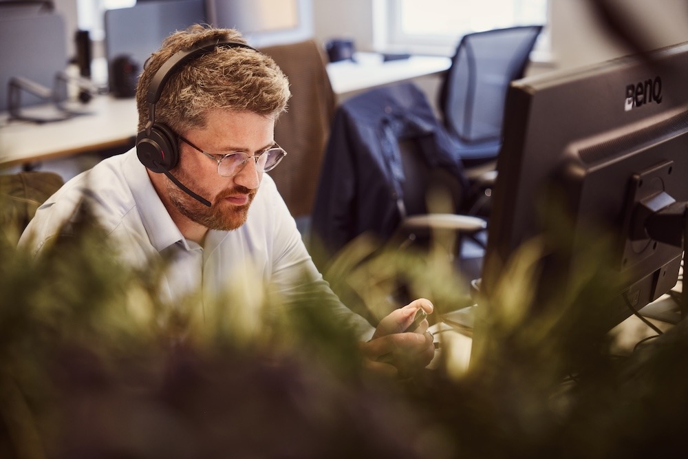 A man wearing glasses and a headset working at his desk in an office environment.