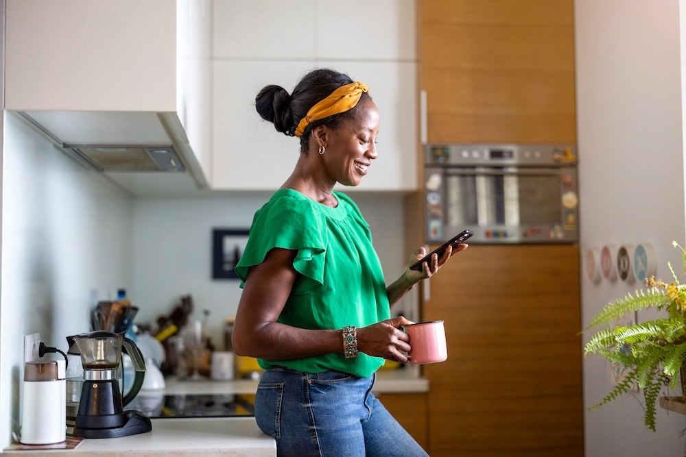 A woman standing in a kitchen holding a mug and looking at a smartphone.