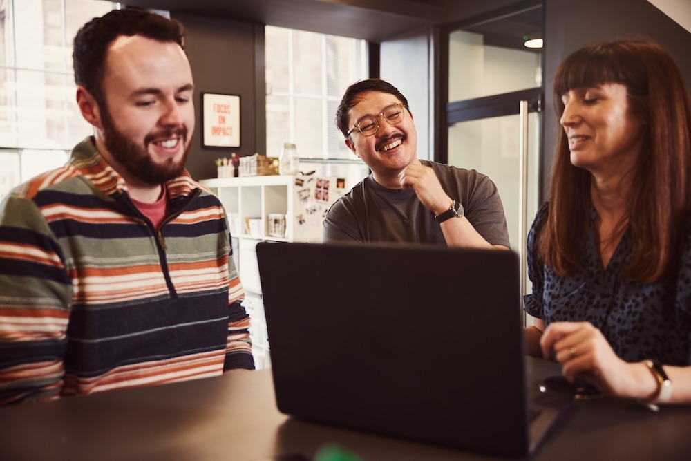 Three colleagues gathered around a table, focused on a laptop and chatting.