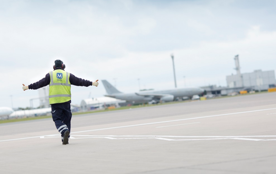 A workman wearing a high vis jacket and headphones walking along an airport tarmac.