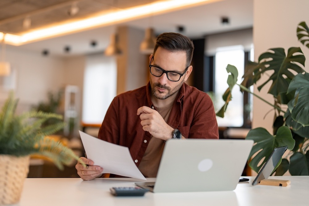 A man wearing glasses studying a piece of paper while sitting at a desk.