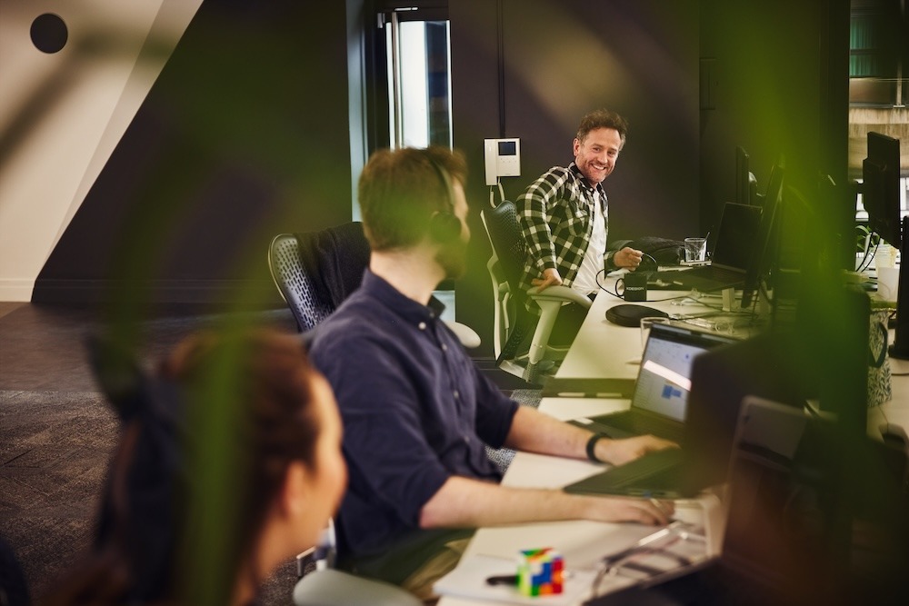 Three colleagues sitting at an island of desks, chatting and smiling.