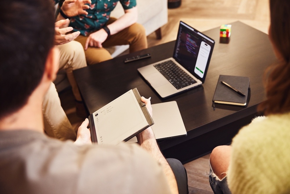 A group of colleagues sitting around a coffee table looking at a laptop. One of them is holding a notebook