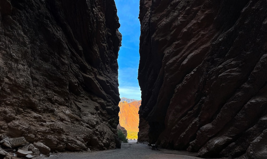 A long, straight path stretches between towering, dark, textured rock walls under a bright blue sky, leading towards a distant, sunlit orange rock formation.