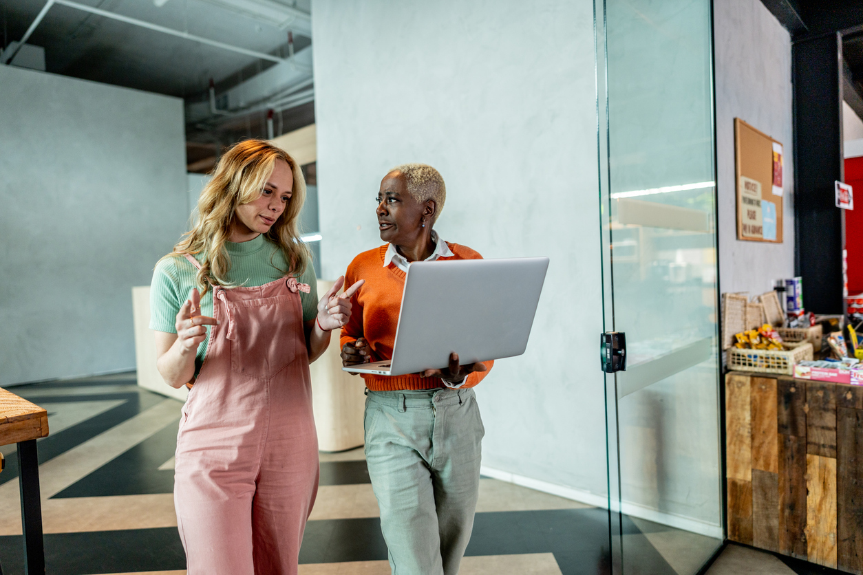 Two colleagues standing in a modern office space, one holding a laptop while the other gestures as they discuss strategy together.